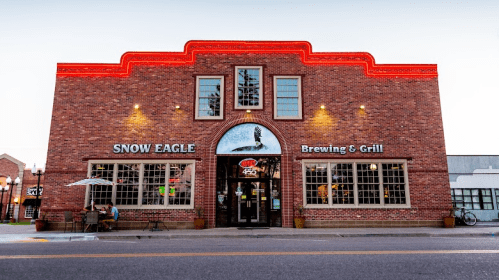 Exterior of Snow Eagle Brewing & Grill, a brick building with large windows and a red-lit roof, on a quiet street.