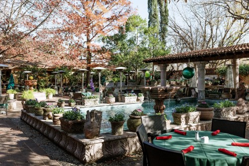 A serene outdoor garden with a fountain, lush greenery, and tables set for dining under sunny skies.