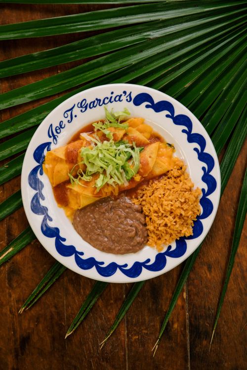 A plate of enchiladas topped with lettuce, served with rice and refried beans, on a decorative blue and white dish.