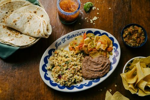 A colorful plate featuring scrambled eggs, refried beans, tortilla chips, and tortillas, with drinks and salsa on the side.