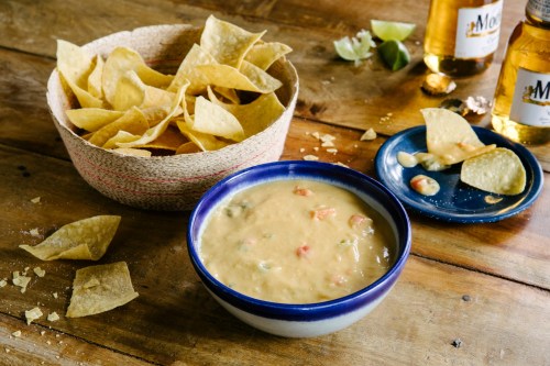 A bowl of creamy dip with diced vegetables, surrounded by tortilla chips and two bottles of beer on a wooden table.
