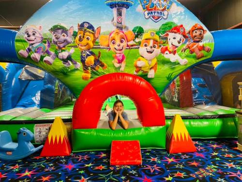 A child peeks out from a colorful bounce house featuring Paw Patrol characters, with vibrant flooring and play equipment around.