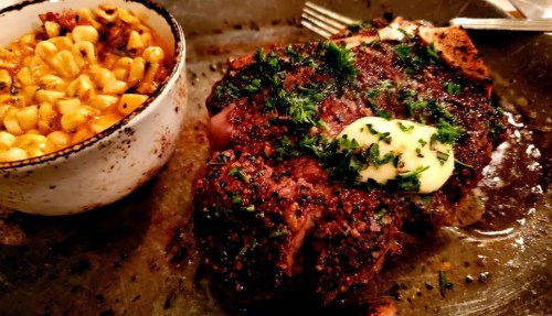 A juicy steak topped with herbs and butter, served alongside a bowl of creamy corn.