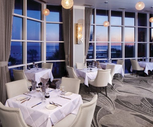 Elegant restaurant interior with white tablecloths, glass windows showcasing a sunset view over the ocean.