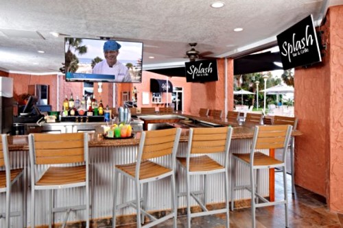 A vibrant bar area with high stools, a TV displaying a chef, and "Splash" signage in a sunny, tropical setting.