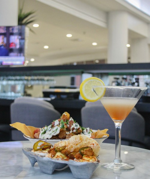 A cocktail with a lemon slice beside a plate of nachos and a dish of crispy snacks on a marble table.