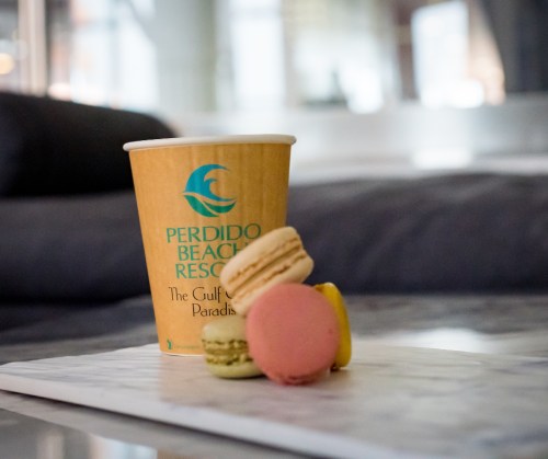 A coffee cup from Perdido Beach Resort next to colorful macarons on a marble surface.