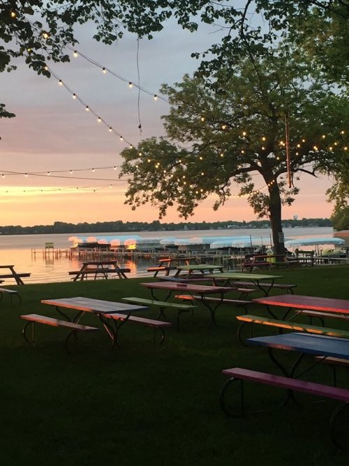 A serene lakeside view at sunset, with picnic tables and string lights illuminating the scene.