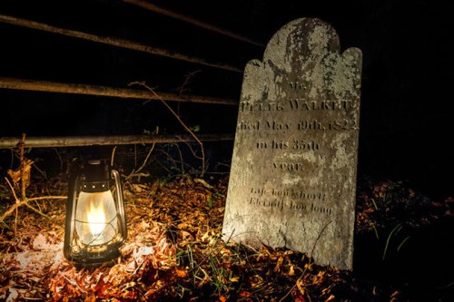 A weathered gravestone beside a lantern, surrounded by fallen leaves in a dark, wooded area.
