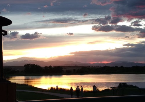 A serene sunset over a lake, with silhouettes of people walking along the shore and mountains in the background.