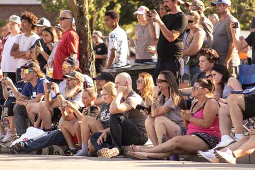 A diverse crowd of people sitting and watching an event, some holding phones, others clapping or engaged.