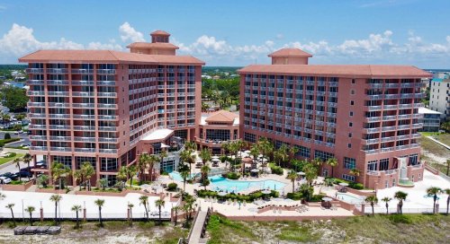 Aerial view of a resort with two large buildings, a pool, and palm trees under a clear blue sky.