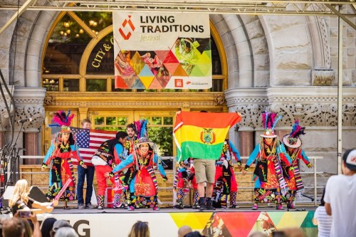 A vibrant cultural performance on stage, featuring dancers in traditional attire holding flags of the USA and Bolivia.