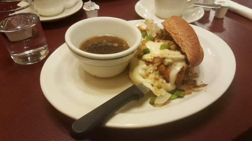 A plate with a sandwich filled with chicken, cheese, and vegetables, alongside a small bowl of dark broth.