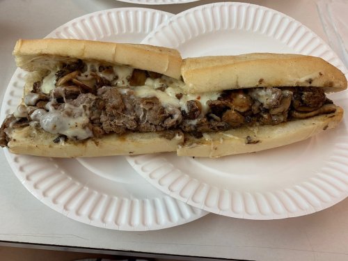 A loaded sandwich with sliced meat, melted cheese, and toppings, served on a white paper plate.