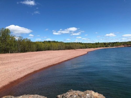 A serene beach with reddish sand, bordered by calm water and green trees under a blue sky with fluffy clouds.