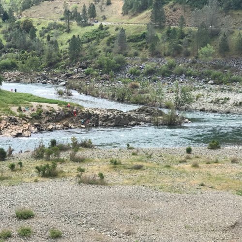 A scenic riverbank with people relaxing on rocks, surrounded by greenery and hills under a clear sky.