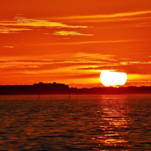 A vibrant sunset over calm waters, with orange and yellow hues reflecting on the surface. Silhouetted land in the background.