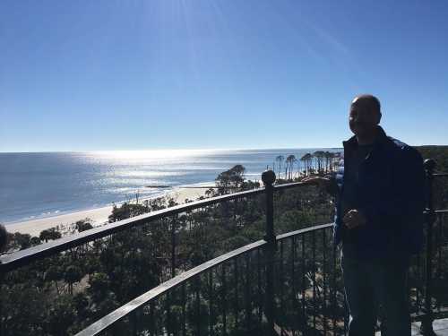 A person stands on a balcony overlooking a sunny beach and ocean, with trees in the background.