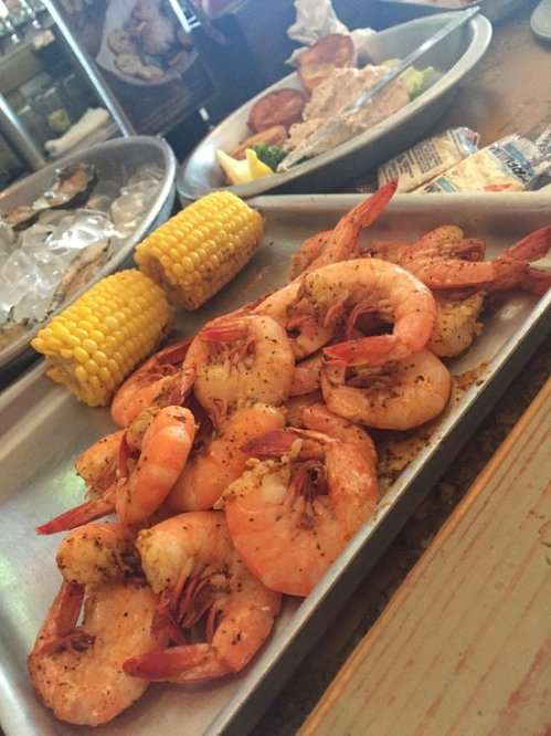 A metal tray filled with seasoned shrimp, corn on the cob, and a side of seafood dishes in the background.