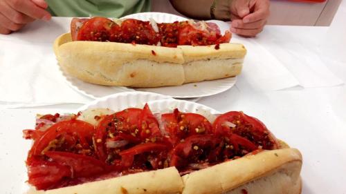 Two large sandwiches on paper plates, filled with sliced tomatoes and a spicy topping, on a white table.