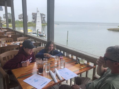 Two children and an adult sit at a wooden table by the water, enjoying drinks and a meal with a scenic view.
