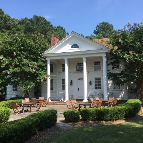 A charming white house with columns, surrounded by greenery and a patio area with wooden chairs and tables.
