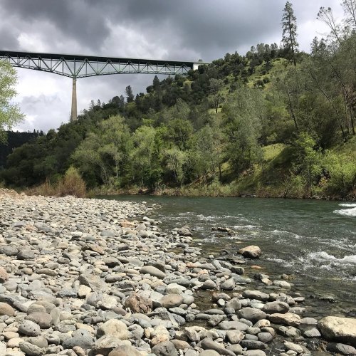 A riverbank with smooth stones, surrounded by lush greenery and a bridge spanning over a valley under cloudy skies.
