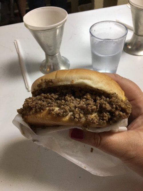 A hand holding a sandwich filled with seasoned ground meat, served on a soft bun, with drinks in the background.