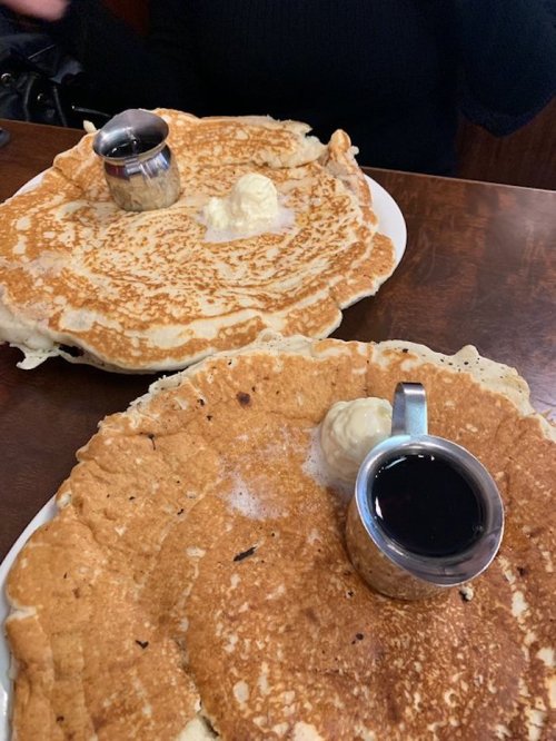 Two large pancakes on plates, each topped with a dollop of butter and served with a small cup of syrup.