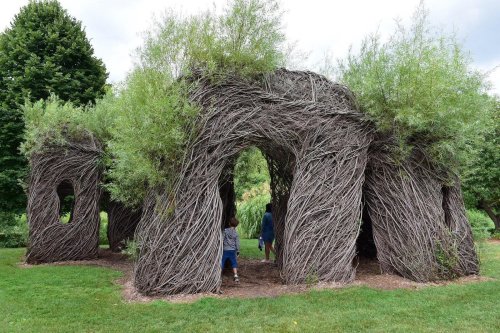 A whimsical structure made of intertwined branches and greenery, with two people walking through it in a park.