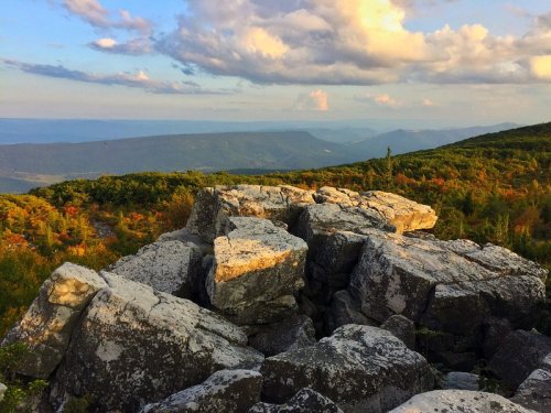 A rocky outcrop overlooks a lush valley and distant mountains under a colorful sky with scattered clouds.