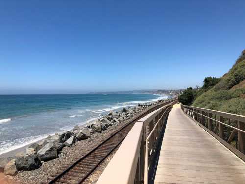 A scenic coastal view with a wooden walkway alongside train tracks and waves lapping at the shore under a clear blue sky.