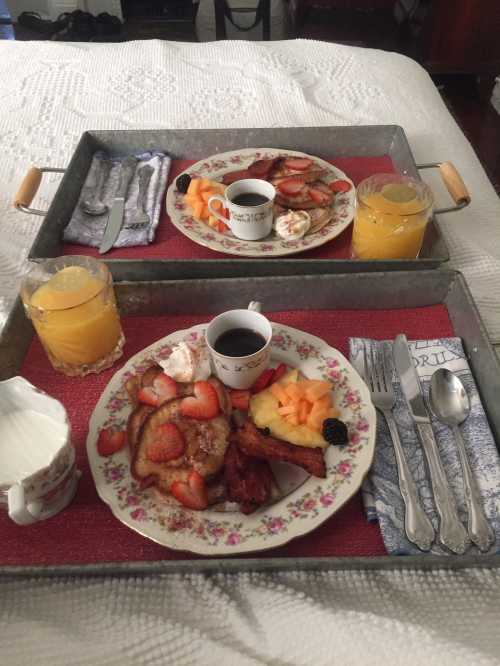 Two trays with breakfast featuring French toast, fruit, bacon, and orange juice, set on a bed with a white quilt.