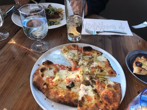 A table setting featuring a pizza with tomatoes and artichokes, a glass of water, and a drink beside a plate of food.