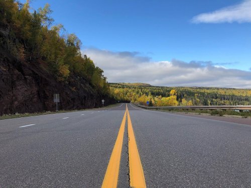 A wide, empty road stretches into the distance, flanked by colorful autumn trees and a cloudy blue sky.