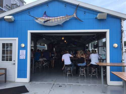 A blue waterfront bar with a marlin decoration, filled with patrons enjoying drinks and socializing inside.