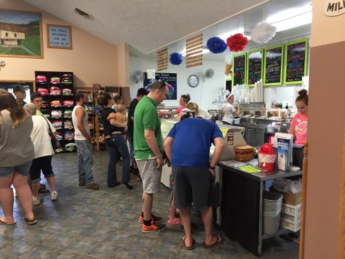 A busy ice cream shop with customers ordering at the counter and others waiting in line. Colorful decorations are visible.
