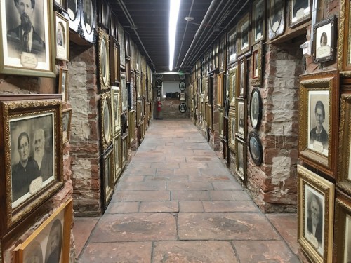 A narrow hallway lined with vintage portraits in ornate frames, set against brick walls and a tiled floor.