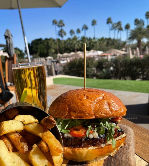 A delicious burger with toppings, served with fries and a beer, against a sunny outdoor backdrop with palm trees.