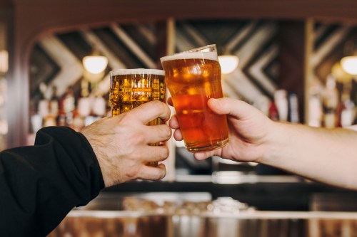 Two hands clink glasses of beer in a bar setting, with a variety of bottles in the background.