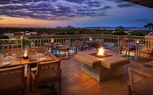 A rooftop dining area with wooden tables, a fire pit, and a scenic sunset view over mountains and palm trees.