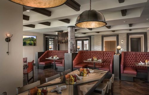 Modern restaurant interior featuring red tufted booths, wooden beams, and a large communal table. Warm lighting enhances ambiance.