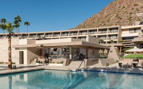 A modern hotel with a pool, bar area, and palm trees, set against a mountain backdrop under a clear blue sky.