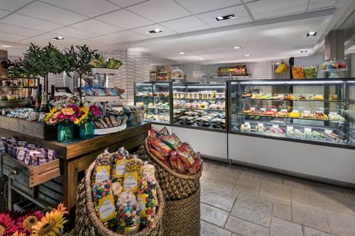 A bright, inviting deli with glass display cases filled with food, colorful flowers, and baskets of snacks.