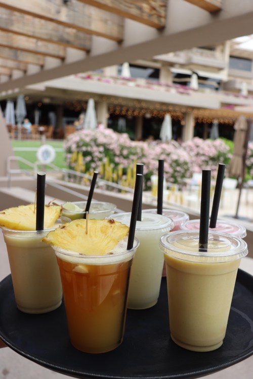 A tray holds several colorful drinks, including pineapple-topped beverages, with a blurred outdoor setting in the background.