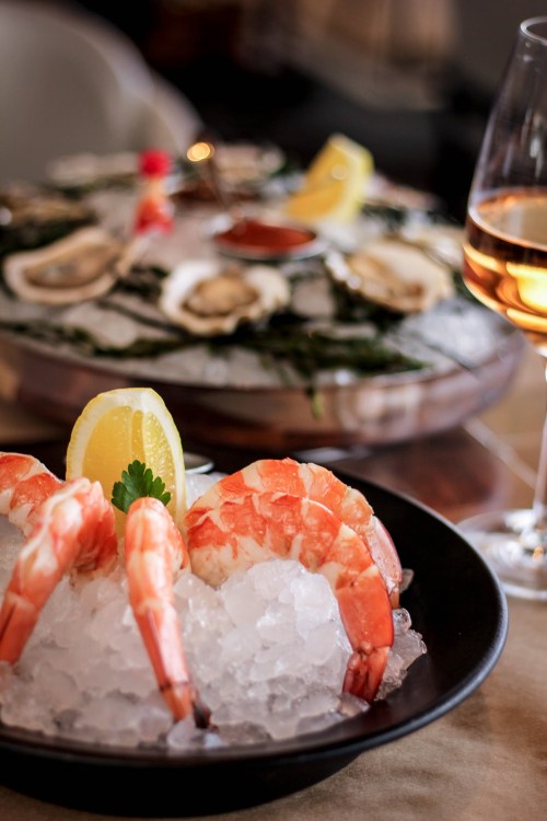 A plate of shrimp on ice with lemon slices, accompanied by a glass of wine and a platter of oysters in the background.
