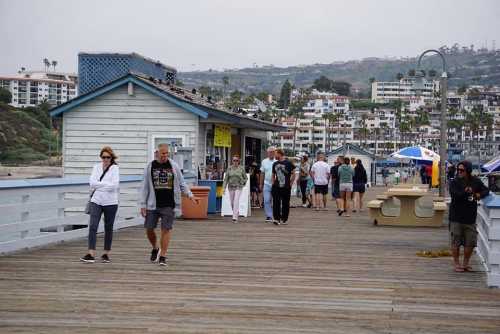 A busy pier with people walking, shops in the background, and a cloudy sky over a coastal town.