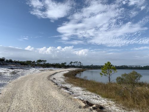 A winding gravel path along a serene lake, surrounded by sandy shores and scattered trees under a blue sky.