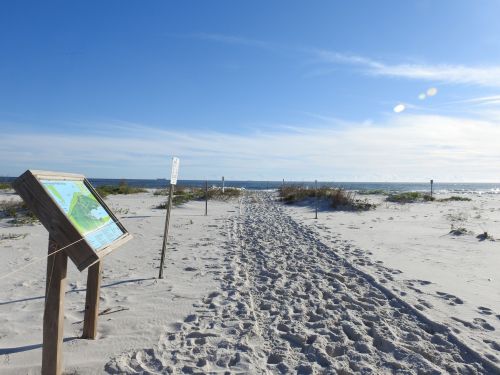 A sandy path leads to the beach, with a map sign on the left and a clear blue sky above.